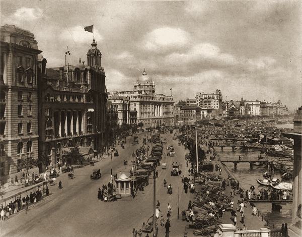 The Bund in Shanghai in the 1920s: a broad waterfront road lined with large stone buildings, busy with cars, rickshaws and people.