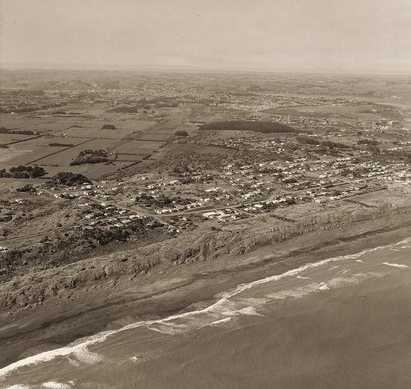 An aerial view of a long beach with houses behind it and farmland beyond.