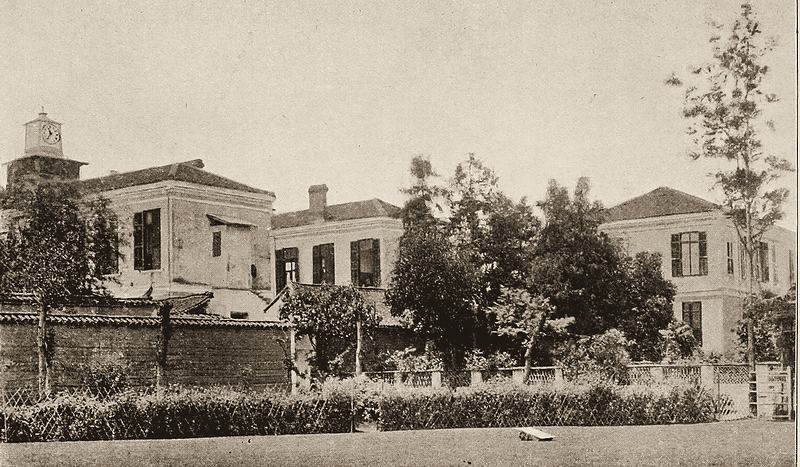Two-storey European-style hospital buildings behind a wall and garden, with a small clock tower.