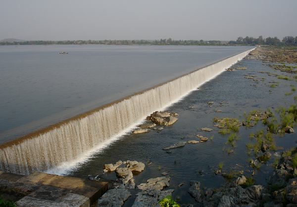Water pouring in a smooth sheet over a long masonry weir, with rocks in the riverbed below.