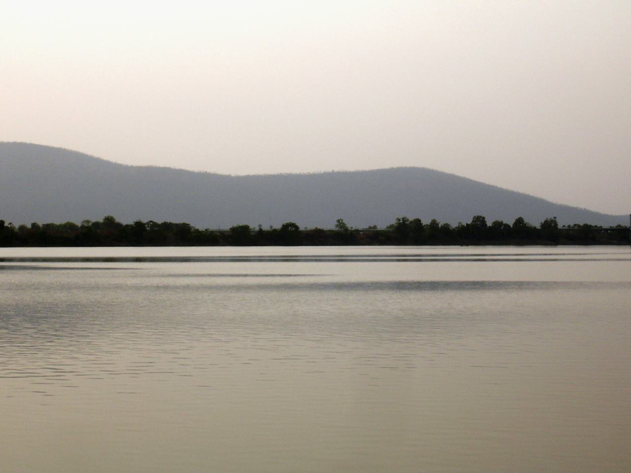 A wide, still river under a hazy sky, with low forested hills on the far bank.