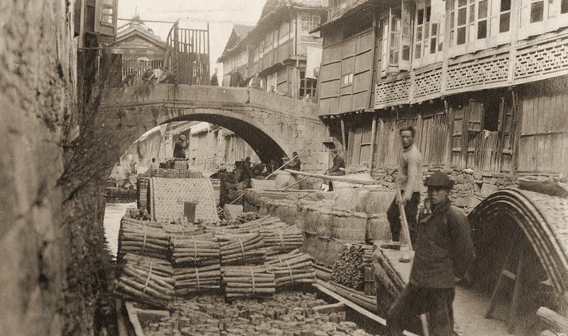 A narrow canal in Hangzhou crossed by a stone arch bridge, lined with wooden houses; a boat piled with bundles of firewood is moored in the foreground.