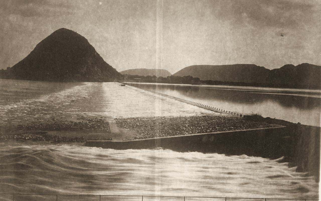 A long stone weir runs across a wide river towards a steep, rounded hill.