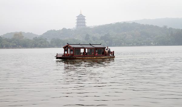 A wooden pleasure boat on misty grey water, with the rebuilt Leifeng Pagoda on a wooded hill behind.