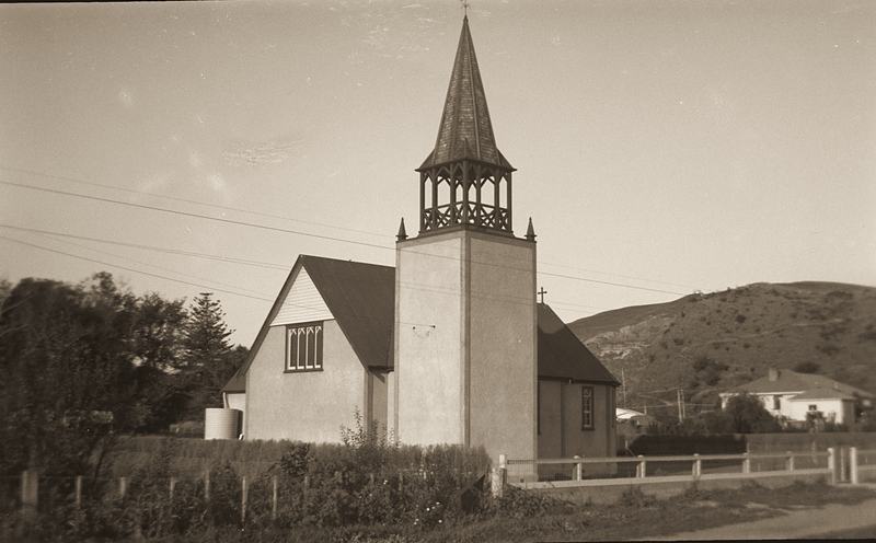 A small white church with a square bell tower topped by an open belfry and spire, with grassy hills behind.