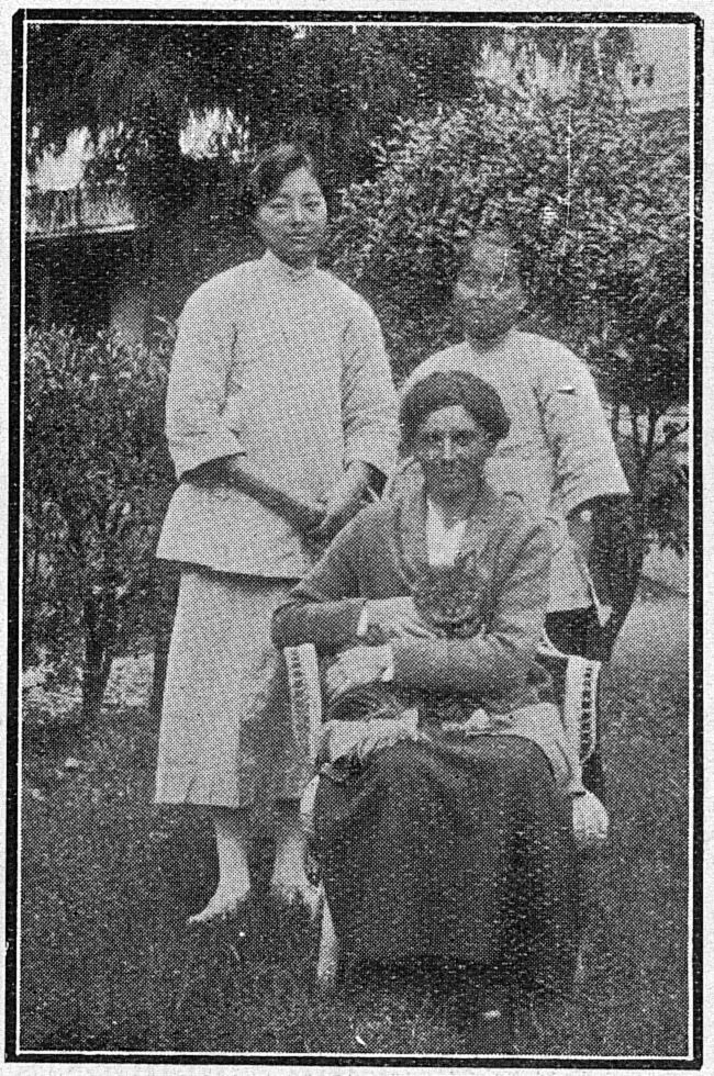 Margaret Woods sits in a wicker chair in a garden with a cat on her lap; two young Chinese women stand behind her.