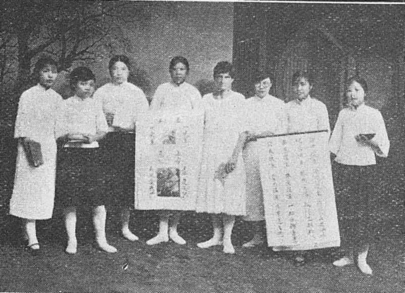 Eight young women in white tunics stand in a row, two of them holding up scrolls of Chinese text.