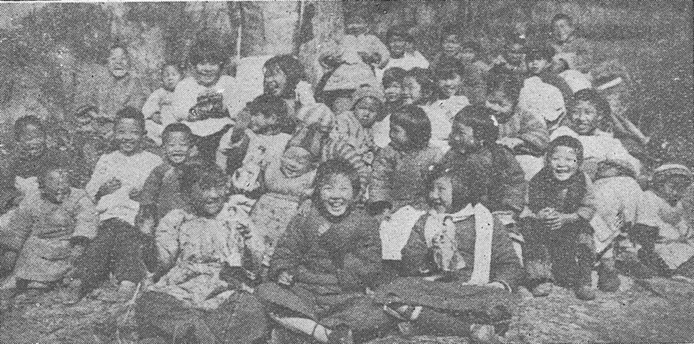 A crowd of small Chinese children in padded winter clothes sit close together on the ground, many of them laughing.