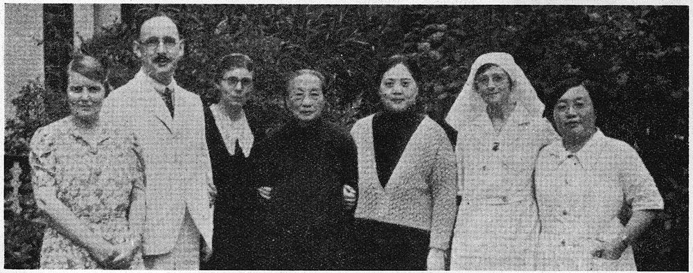 Seven people stand in a row outside: Western and Chinese doctors, wives and nurses, one nurse in a white veil.