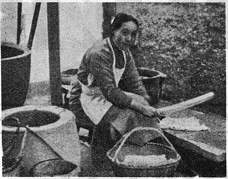 A woman in an apron sits beside large stone jars, working with a wooden paddle over a basket.