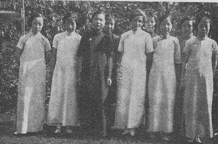 A row of young Chinese women in long qipao dresses standing in front of trees.