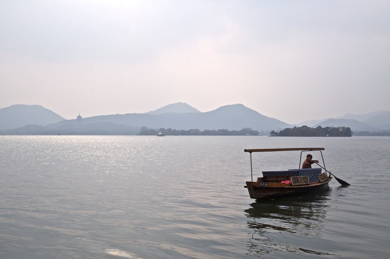 A lone boatman sculling a small covered boat across West Lake, with layers of misty hills behind.