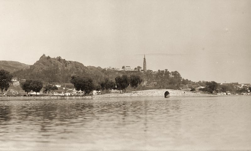 Across the water to a rocky hill with a slender pagoda on top and an arched bridge at its foot.