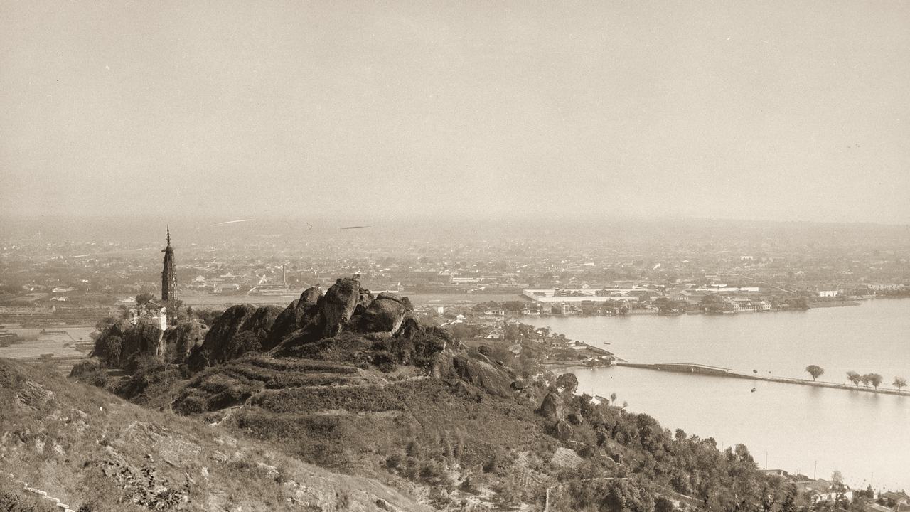 Looking down over a slender pagoda on a rocky hill to a causeway across West Lake and the flat roofs of Hangzhou beyond.