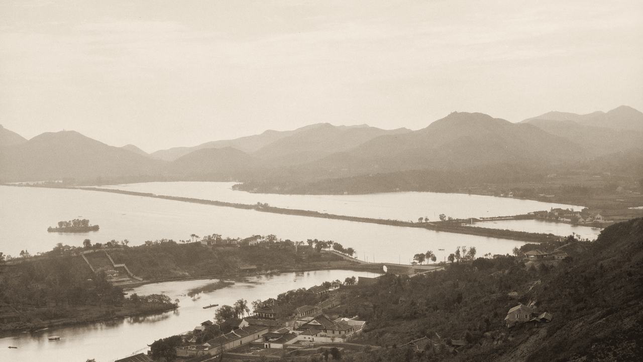 West Lake at Hangzhou in the early 1920s, seen from the hills: a long causeway crosses the water towards a range of mountains.