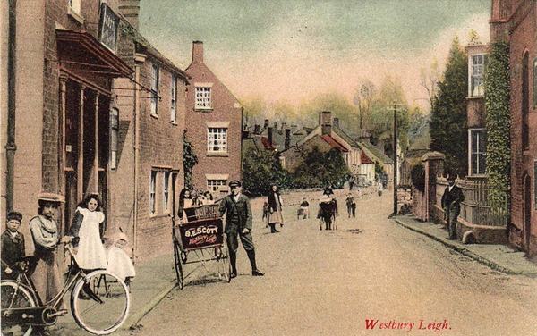 A hand-coloured postcard of a village street of brick houses, with children, a man with a handcart and a bicycle.